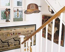 Decorating the lower stair landing is an 1887 Lancaster, Pennsylvania, hooked rug from Olde Hope Antiques, and a 1910 rug with a horse and carriage scene from Jeff Bridgman. The 19th-century hat trade sign from the Dr. and Mrs. Ed Hoffman collection was purchased at Northeast Auctions, Portsmouth, New Hampshire. On the window sill is a 20th-century figure by outsider artist William Duffy (left), and an 1880s figure of a man slicing a watermelon.