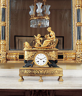 A French nineteenth-century mantel clock probably designed for export, is adorned with a goddess and putti on a chariot pulled by butterflies. A Messenger and Sons chandelier, 1825-1830, is reflected in the parcel-gilt and ebonized mirror, Boston, 1915.