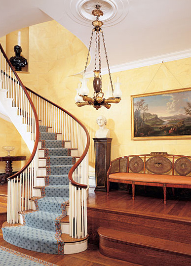 Many of the home's lighting fixtures were most likely purchased by the original owners. This chandelier is from Thomas Messenger and Sons of London and Birmingham, England, circa 1830. As one of the largest makers of oil lamps in England at the time, the Messenger firm provided merchandise to retailers from Boston to New Orleans. The raised platform visible here was built to provide enough headroom to walk horses through a passageway below that ran from a preexisting stable on another property to the street. When the couple moved in, the stair railings were covered in a black paint, which the couple removed. The classical landscape painting is by Michel Felice Cornè (1752-1845). Cornè emigrated from Italy to America as a young man. Before moving to Boston in 1806, he helped establish the tradition of marine painting in the Salem area.